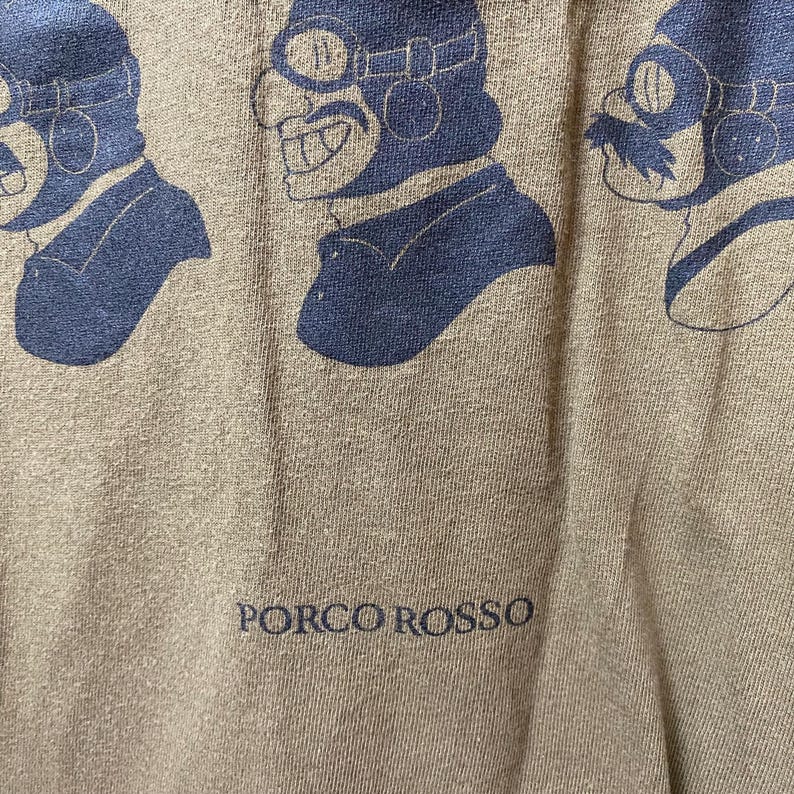 May include: Close-up of a tan t-shirt featuring a repeating blue graphic of a character wearing goggles and a pilot's cap. The text "PORCO ROSSO" is printed in blue at the bottom of the shirt.
