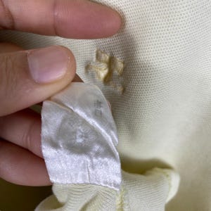May include: Close-up of a light yellow polo shirt with a small hole near the collar. A white tag with Japanese text is held up, showing the garment's label. The fabric has a textured appearance.