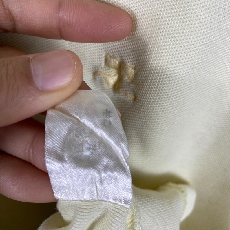 May include: Close-up of a light yellow polo shirt with a small hole near the collar. A white tag with Japanese text is held up, showing the garment's label. The fabric has a textured appearance.