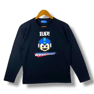 May include: Black long-sleeve shirt featuring a pixelated graphic of a blue-helmeted character with the text "1UP!" above and "ROCKMAN" below in red and blue. The shirt is hanging on a wooden hanger.