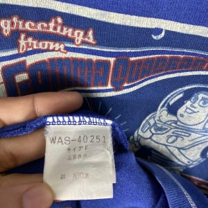 May include: Blue t-shirt with a white and blue graphic print of a cartoon character. The shirt has a white tag with the text "WAS-40251", "サイズ F", "品質表示", and "綿 100%".