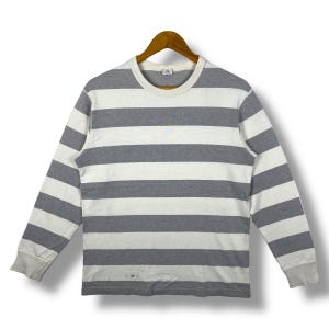 May include: A long-sleeved striped shirt in grey and white, hanging on a wooden hanger. The shirt features a crew neck and ribbed cuffs. The horizontal stripes are evenly spaced. The garment is displayed against a white backdrop, suggesting a studio setting.