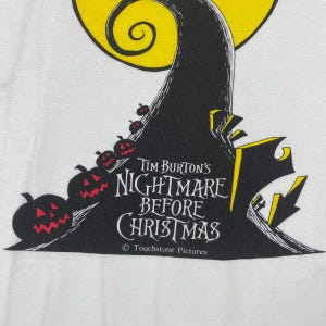 Puede incluir: Camiseta blanca con una ilustraci&oacute;n en negro y amarillo de Jack Skellington de "Pesadilla antes de Navidad". La ilustraci&oacute;n incluye una luna amarilla, un &aacute;rbol negro y una l&iacute;nea de calabazas rojas y negras. El texto "Tim Burton's Nightmare Before Christmas" y "Touchstone Pictures" est&aacute; impreso en negro.