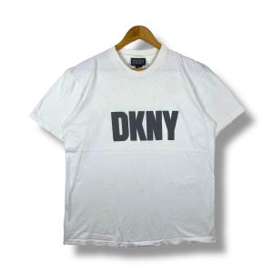 May include: White DKNY t-shirt with a large, dark grey DKNY logo across the chest. The shirt is hanging on a wooden hanger against a white background. The DKNY logo is centered and bold.