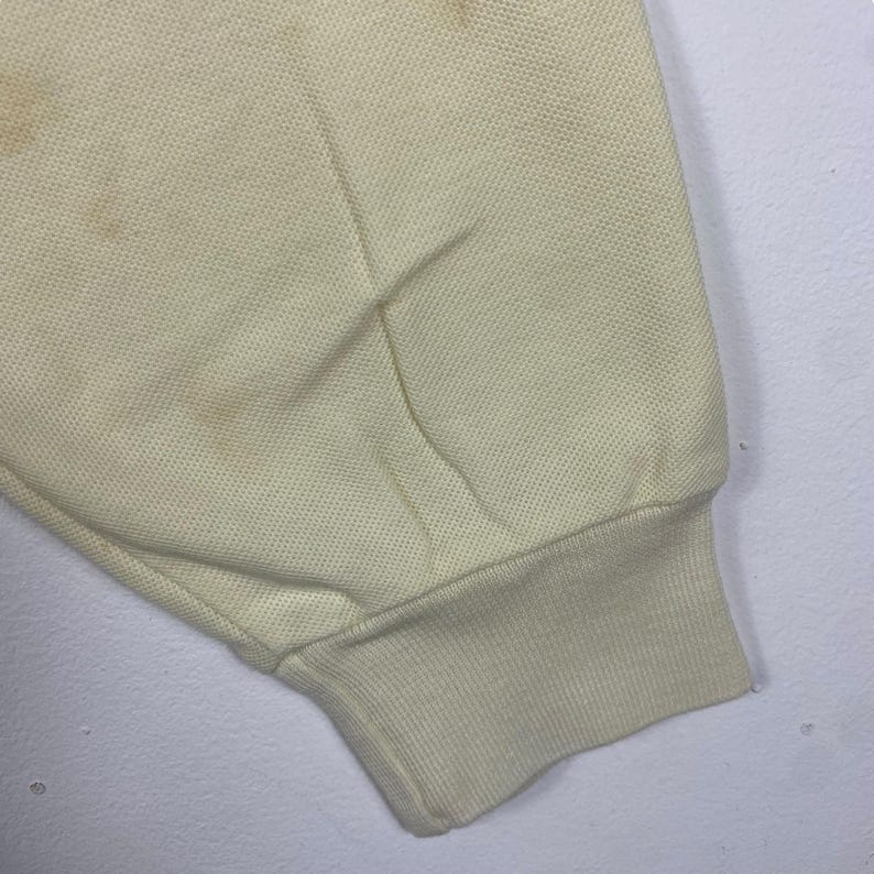 May include: Close-up of a pale yellow fabric sleeve with a ribbed cuff. The fabric has a textured appearance and shows several brown stains. The sleeve is slightly wrinkled.