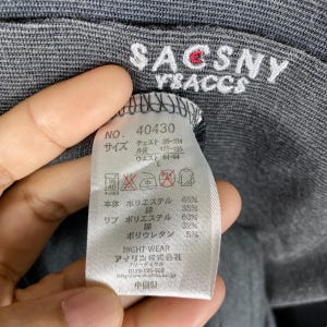K&ouml;nnte beinhalten: Ein graues Langarmshirt mit dem Aufdruck "SACSNY YSACCS" auf der Vorderseite. Das Shirt hat ein Etikett mit dem Text "NO. 40430" und Gr&ouml;&szlig;enangaben in Japanisch. Das Etikett listet auch die Stoffzusammensetzung auf: 65 % Polyester, 35 % Baumwolle f&uuml;r den K&ouml;rper und 63 % Polyester, 32 % Baumwolle, 5 % Polyurethan f&uuml;r die Rippen.