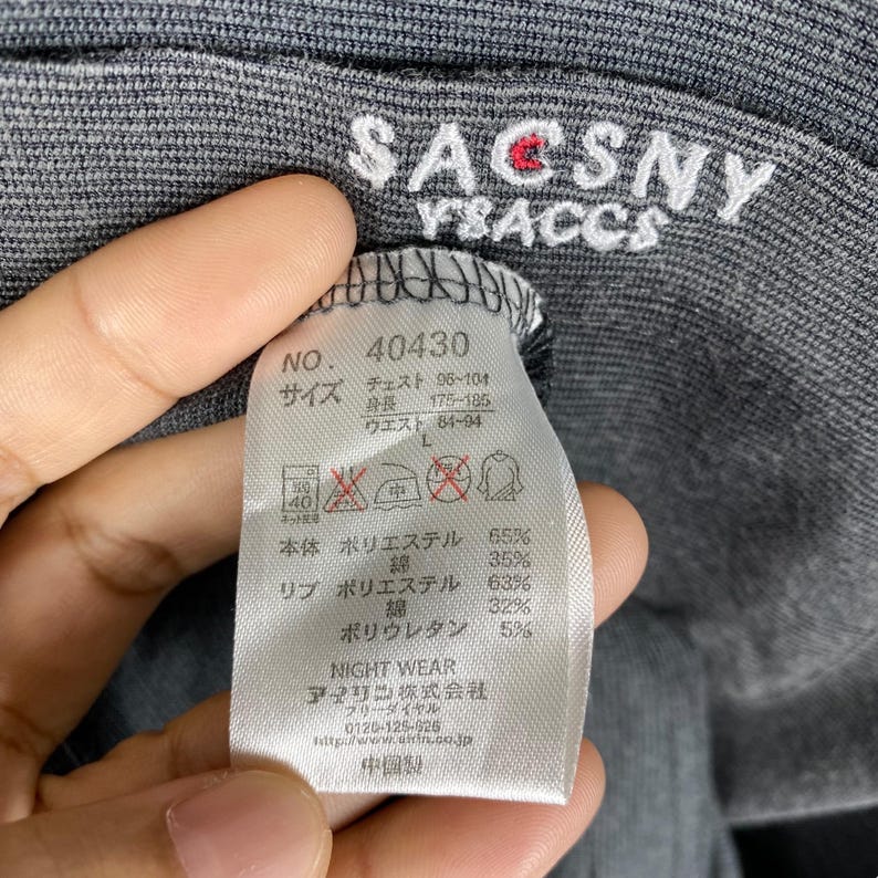 K&ouml;nnte beinhalten: Ein graues Langarmshirt mit dem Aufdruck "SACSNY YSACCS" auf der Vorderseite. Das Shirt hat ein Etikett mit dem Text "NO. 40430" und Gr&ouml;&szlig;enangaben in Japanisch. Das Etikett listet auch die Stoffzusammensetzung auf: 65 % Polyester, 35 % Baumwolle f&uuml;r den K&ouml;rper und 63 % Polyester, 32 % Baumwolle, 5 % Polyurethan f&uuml;r die Rippen.
