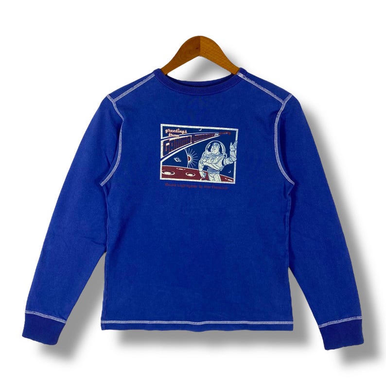 May include: A blue long-sleeved t-shirt with a white stitched trim. The shirt features a graphic of Buzz Lightyear from the Toy Story franchise with the text "Greetings from Galactic Command".