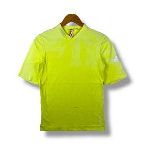 Könnte beinhalten: Ein gelbes T-Shirt mit kurzen Ärmeln und V-Ausschnitt mit einem weißen Logo auf dem rechten Ärmel. Das Shirt hat einen verblassten Tie-Dye-Effekt.