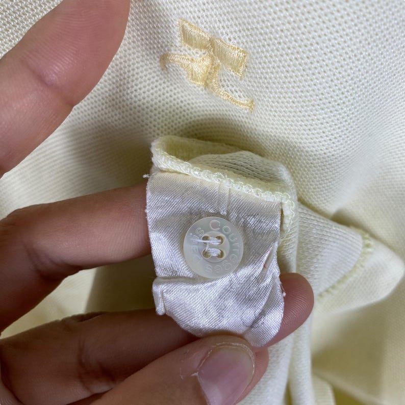 May include: Close-up of a pale yellow polo shirt collar with a small embroidered bow detail. The shirt's label is visible, featuring a button and printed text. The fabric appears to be a soft knit.