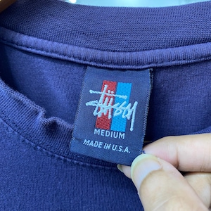 May include: Close-up of a navy blue t-shirt with a tag featuring a logo in red and blue. The tag reads "MEDIUM" and "MADE IN U.S.A." in white text. The shirt's fabric is visible, showing its texture.