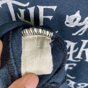 Puede incluir: Primer plano de una camiseta azul marino con texto blanco y un gr&aacute;fico de gato. El texto incluye la palabra "THE". Una etiqueta blanca es visible dentro del cuello de la camisa.