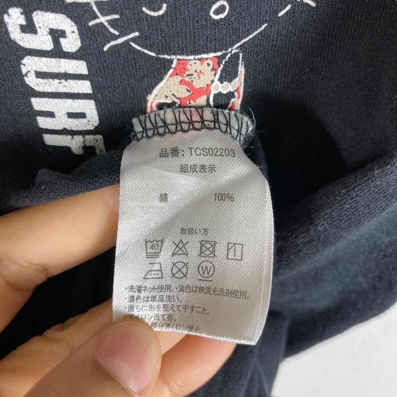 Pode incluir: Close-up de uma camiseta preta com a palavra "SURF" em branco. Uma etiqueta com texto japon&ecirc;s e s&iacute;mbolos de cuidados est&aacute; anexada. A etiqueta inclui o texto "100%" e "綿".