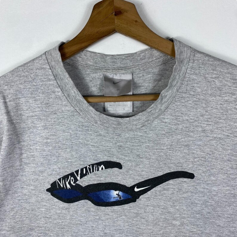 Może przedstawiać: Szary T-shirt z logo Nike i nadrukiem "Nike Vision" z przodu. Tekst jest biały i otoczony czarną i niebieską grafiką okular&oacute;w przeciwsłonecznych.