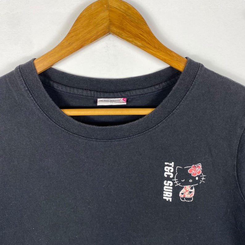 Pode incluir: Camiseta preta de gola redonda com um pequeno gr&aacute;fico de um gato com uma flor e o texto "TGC SURF" em branco. A camisa est&aacute; pendurada em um cabide de madeira.