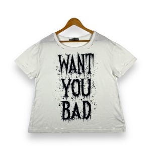 Può includere: Maglietta bianca con un aspetto consumato e una stampa nera che dice "WANT YOU BAD".