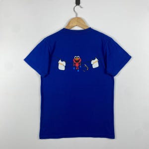 May include: A blue t-shirt featuring a graphic of Elmo hanging from a clothesline with two white shirts. The text on the shirts reads "Elmo".
