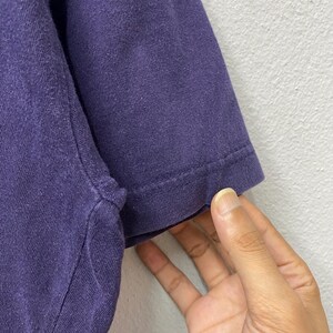 May include: Close-up of a section of a dark purple t-shirt sleeve. The sleeve is made of a soft, textured fabric. The edge of the sleeve is hemmed with a visible seam. A hand is touching the sleeve.