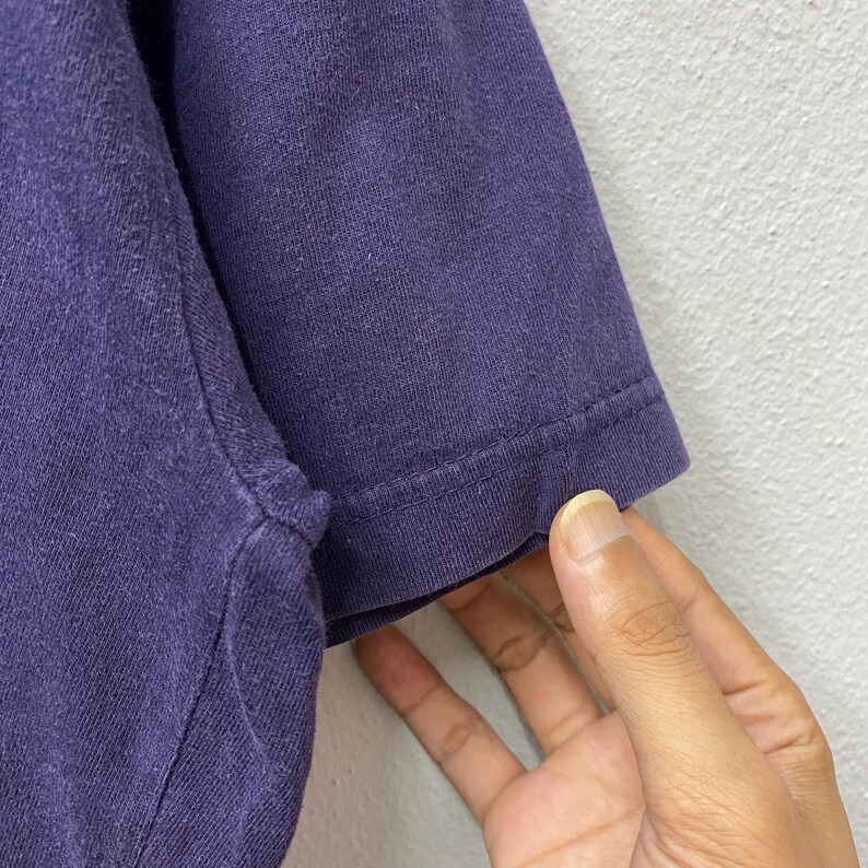 May include: Close-up of a section of a dark purple t-shirt sleeve. The sleeve is made of a soft, textured fabric. The edge of the sleeve is hemmed with a visible seam. A hand is touching the sleeve.