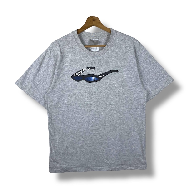 Może przedstawiać: Szara koszulka z kr&oacute;tkim rękawem z grafiką przedstawiającą okulary przeciwsłoneczne z napisem "Nike Vision" na soczewce.