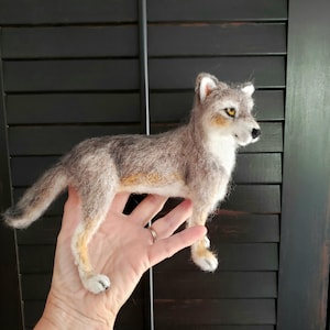 May include: A needle-felted wolf figurine featuring a coat of blended gray, white, and tan fibers. The wolf is depicted in a standing position with alert ears, amber-colored eyes, and a black nose, showcasing detailed texture throughout its body.