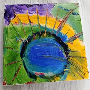 Puede incluir: Pintura abstracta con un círculo azul rodeado de pintura amarilla, verde y morada. El círculo tiene un contorno negro y está rodeado de finas líneas puntiagudas de pintura roja y amarilla.