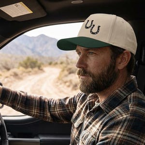 May include: A beige baseball cap with a dark green brim and a black horseshoe logo is worn by a person driving a vehicle. The person is wearing a plaid shirt. The background shows a dirt road and mountains.