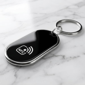 Peut inclure: Un porte-clés NFC noir avec une bordure argentée et un porte-clés argenté. Le porte-clés a une finition brillante et présente le symbole NFC en blanc. Le fond est une surface en marbre blanc.