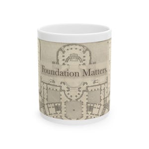 May include: White ceramic mug featuring a detailed architectural blueprint design in beige and gray tones. The mug has the text "Foundation Matters" printed on it. The mug is cylindrical and has a white interior.