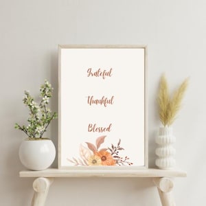Op de afbeelding: Een printbare wandkunstprint met de woorden "Grateful", "Thankful" en "Blessed" in een koperkleurige lettertype. De print heeft een aquarelbloemendesign in tinten oranje, perzik en bruin.