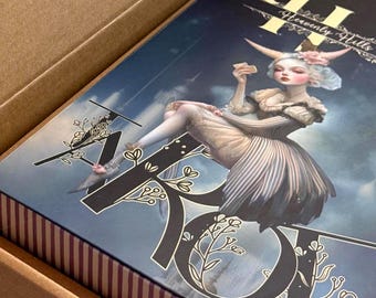 Puede incluir: Una caja decorativa con una ilustración caprichosa de una figura sentada con cuernos, sosteniendo una carta. La caja está dentro de un contenedor de cartón. El texto en la caja dice "Heavenly Hills". El interior de la tapa de la caja tiene el texto "Hello, my dear!"