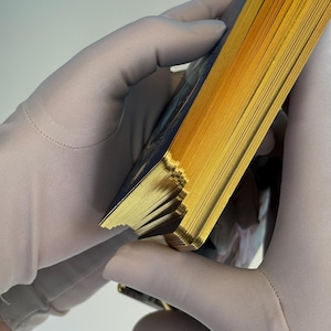 May include: A deck of cards with gold gilded edges, held by gloved hands. The cards have a dark blue design on the back. The edges of the cards are a bright gold color.