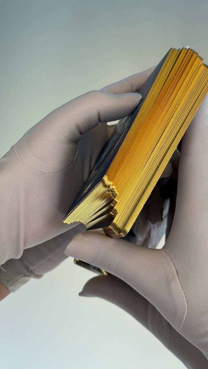 May include: A deck of cards with gold gilded edges, held by gloved hands. The cards have a dark blue design on the back. The edges of the cards are a bright gold color.