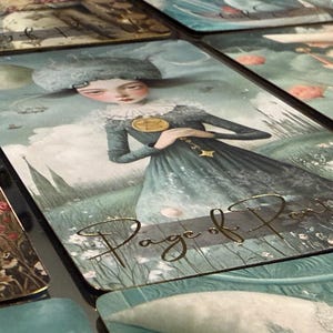 May include: Tarot card featuring a woman in a blue dress with a gold pendant, standing in a field. The card is labeled "Page of Pentacles" in gold script. Other cards with similar artwork are visible.