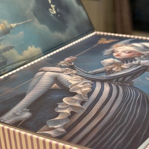 May include: Decorative box with a whimsical illustration. The lid features a seated figure in a striped dress, holding a staff, with a crescent moon and text. The box has a striped pattern on the sides and a sky scene on the inside of the lid.