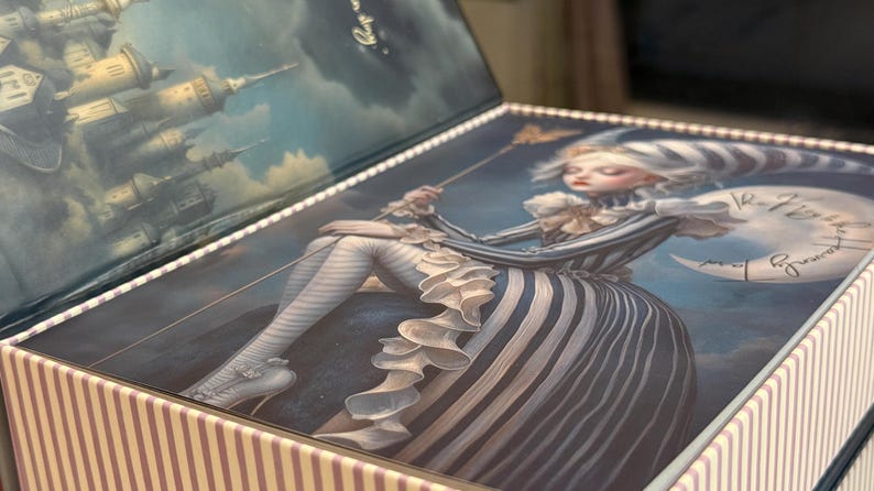 May include: Decorative box with a whimsical illustration. The lid features a seated figure in a striped dress, holding a staff, with a crescent moon and text. The box has a striped pattern on the sides and a sky scene on the inside of the lid.