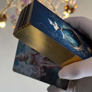 May include: A deck of tarot cards with gold gilded edges. The cards feature a blue and white illustration of a celestial scene. The edges of the cards are a bright gold color. The cards are held in a gloved hand.
