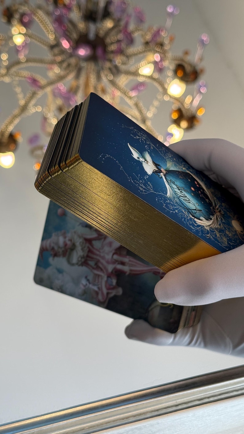 May include: A deck of tarot cards with gold gilded edges. The cards feature a blue and white illustration of a celestial scene. The edges of the cards are a bright gold color. The cards are held in a gloved hand.