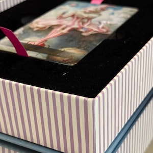 May include: A decorative box with a striped pattern in white and lavender. Inside, a black velvet lining holds a rectangular card with a painting of a ballerina. A pink ribbon is attached to the card.