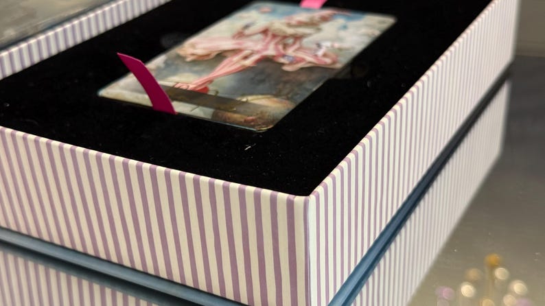 May include: A decorative box with a striped pattern in white and lavender. Inside, a black velvet lining holds a rectangular card with a painting of a ballerina. A pink ribbon is attached to the card.