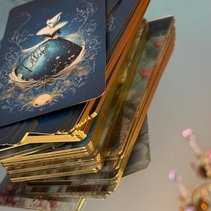 May include: A deck of tarot cards with a blue and gold design featuring a woman in a dress. The cards have gold edges and are stacked on top of each other. The background is blurred.