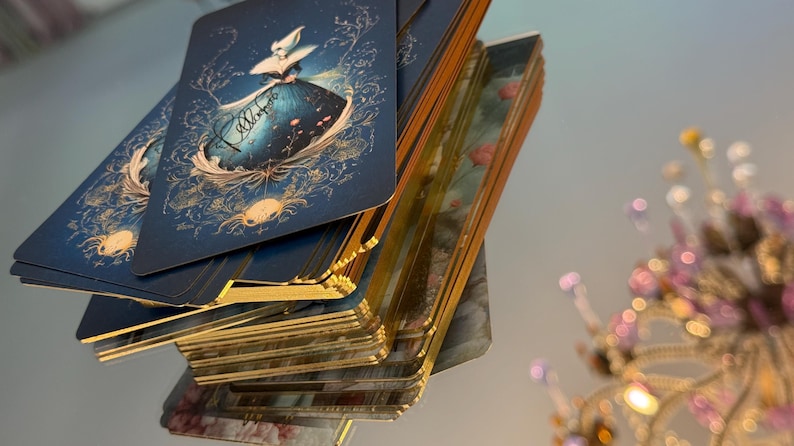 May include: A deck of tarot cards with a blue and gold design featuring a woman in a dress. The cards have gold edges and are stacked on top of each other. The background is blurred.