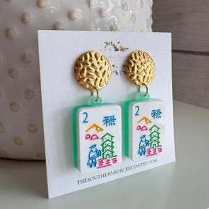 May include: Gold-coloured stud earrings with a textured surface, connected to white and green rectangular charms. The charms feature colourful illustrations of Chinese characters and symbols, with the number "2" in blue. The earrings are displayed on a white card.