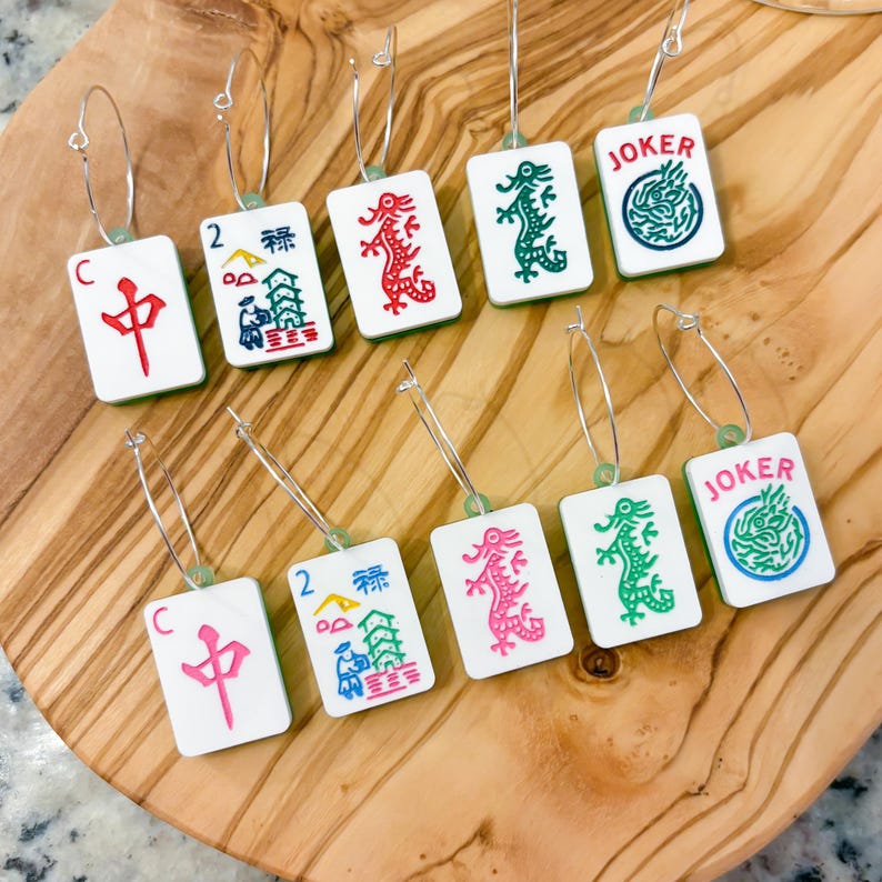 May include: A collection of white rectangular earrings with various colorful designs, including dragons, Chinese characters, and the word "JOKER". Each earring is attached to a silver hoop. The earrings are displayed on a wooden cutting board.