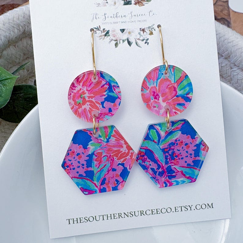 May include: A pair of dangle earrings with a floral pattern in pink, blue, and green. The earrings have a gold-colored hook and are made of two geometric shapes: a circle and a hexagon. The text "The Southern Surcee Co." and the Etsy shop URL are visible.