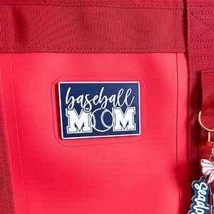 May include: A red tote bag with a white and navy blue "baseball MOM" sticker. The sticker features a baseball graphic. The bag has red straps and a red handle. A keychain with a baseball theme is attached to the bag.