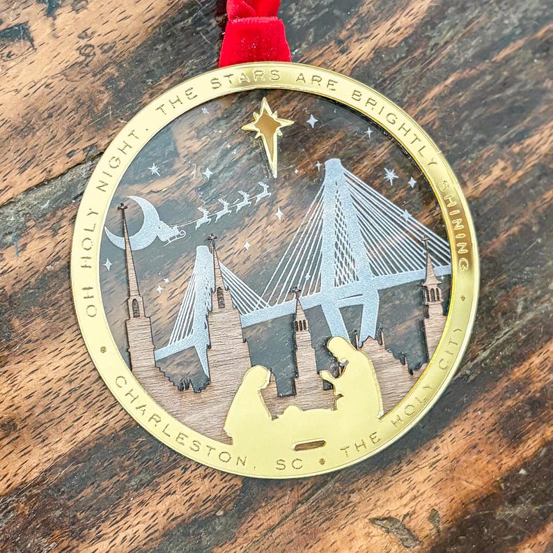 May include: A gold-colored Christmas ornament with a red ribbon. The ornament features a laser-cut design of a bridge, buildings, and a nativity scene. Text around the edge reads "OH HOLY NIGHT. THE STARS ARE BRIGHTLY SHINING CHARLESTON, SC. THE HOLY CITY."