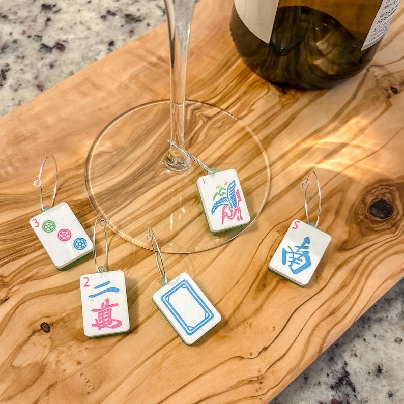 May include: A collection of Mahjong tile wine charms. Each charm features a different tile design with various colors and symbols. The charms are attached to silver hoops and are displayed on a wooden cutting board.