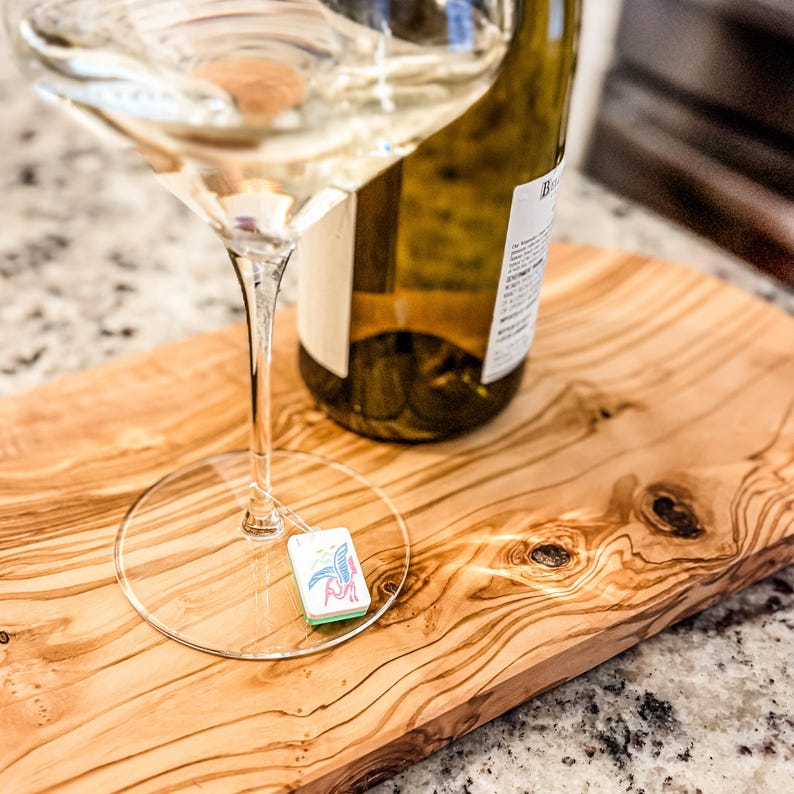 May include: A wine glass with a clear stem and a small tag with a colorful design rests on a wooden cutting board. A partially visible wine bottle sits behind the glass. The board has a natural wood grain.