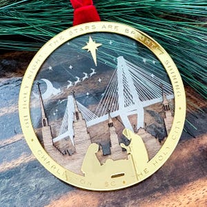 May include: A gold-colored Christmas ornament with a laser-cut design featuring a bridge, church steeples, and a nativity scene. The ornament has the words "OH HOLY NIGHT. THE STARS ARE BRIGHT! SHINING. CHARLESTON SC. THE HOLY CITY" around the edge.